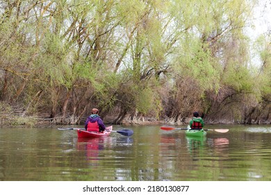 Rear view of two women sitting in red and green kayaks. Spring Danube river near trees with gentle green leaves