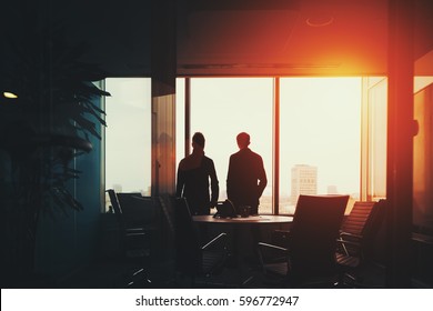 Rear View Of Two Young Businessmen Near Big Window And Work Table With Many Armchairs In Dark Office Having Rest, Discussing Something And Looking Outside After Meeting, Metropolitan Cityscape Outside