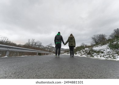 Rear view of two young people in the middle of the road. A couple holding hands while walking along a road in a snowy mountain landscape under a cloudy sky. - Powered by Shutterstock