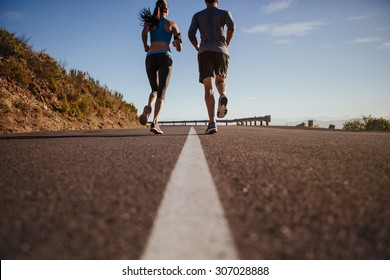 Rear View Of Two Young People Training Together On Road. Man And Woman On Morning Run On Summer Day. Low Angle Shot Of Couple Jogging Outdoors.