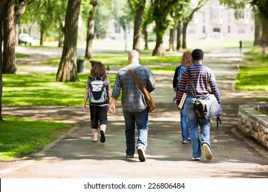 Rear View Of University Students With Backpacks Walking On Campus Road