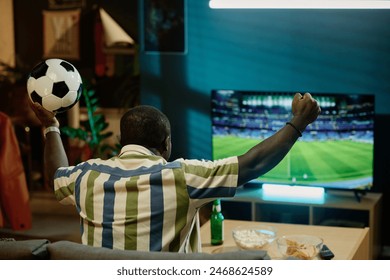 Rear view of unrecognizable African American soccer fan holding ball watching match on TV in living room at home, copy space - Powered by Shutterstock