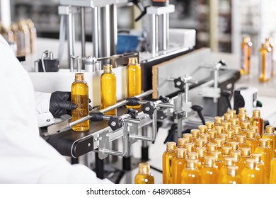 Rear view of unrecognizable factory worker in gown and gloves controlling production process of manufacturing cosmetics at modern laboratory, taking bottle of golden yellow liquid from conveyor line