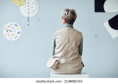 Rear View Of Unrecognizable Mature Woman Sitting On Stool In Modern Art Gallery Looking At Artworks