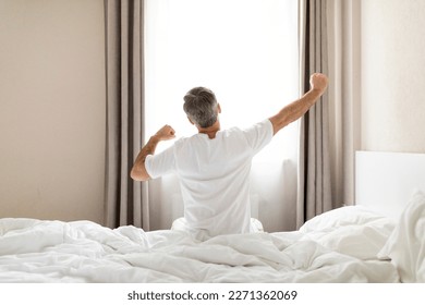 Rear view of unrecognizable middle aged man in pajamas sitting on bed and stretching body after waking up in the morning, looking at window, copy space. Comfortable healthy sleep concept - Powered by Shutterstock