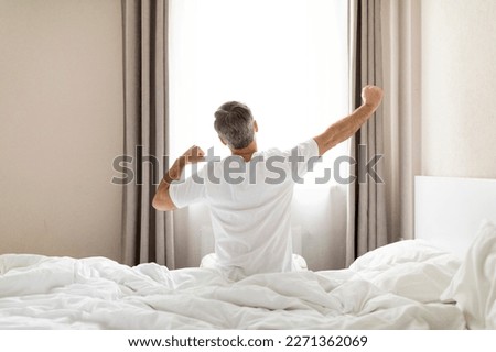 Rear view of unrecognizable middle aged man in pajamas sitting on bed and stretching body after waking up in the morning, looking at window, copy space. Comfortable healthy sleep concept