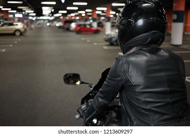 Rear View Of Unrecognizable Woman Wearing Stylish Motorcycle Leather Jacket, Gloves And Safety Helmet, Sitting In Her Two Wheeled Motor Vehicle In Underground Garage Or Car Parking, Ready To Drive