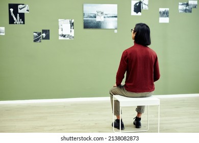 Rear View Of Unrecognizable Young Man Sitting On Stool In Modern Art Gallery Looking At Black And White Photos On Green Wall