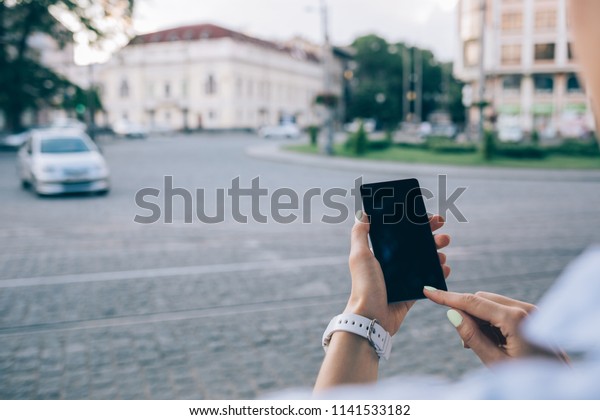 Rear view unrecognizable young woman using smart\
phone standing near european paving stone road with cars. Close-up\
female hands touching device screen on the background of old city\
street.
