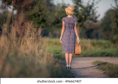 Rear view of vintage 1920s summer fashion woman with blue dress and straw hat standing with handbag in rural landscape.