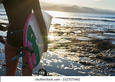 Rear View Of A Wet Suit Clad Female Surfer Holding A Surf Board Near The Waters Edge