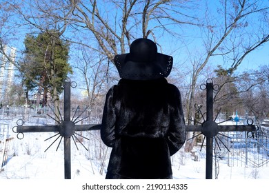 Rear View Of A Woman In A Black Fur Coat And A Hat In A Cemetery In Winter. Woman Between Two Black Crosses. Graves In Winter. Old Cemetery