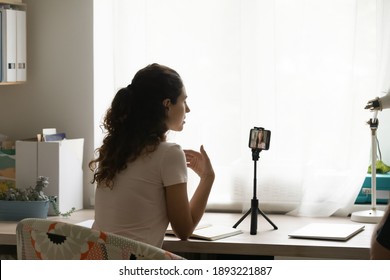 Rear view woman blogger recording vlog on smartphone on tripod, young female vlogger shooting video for social network, webinar or online training, speaking, sitting at work desk at home
