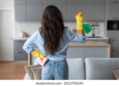 Rear view of woman in blue shirt and denim jeans, wearing yellow gloves, holding spray bottle and rag while cleaning her modern kitchen - Powered by Shutterstock