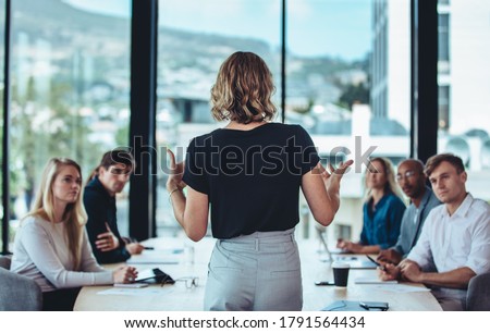 Rear view of a woman explaining new strategies to coworkers during conference meeting in office. Businesspeople meeting in office board room for new project discussion.