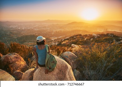 Rear view of woman hiker sitting on rock on top of hill while looking at sunset over San Diego California