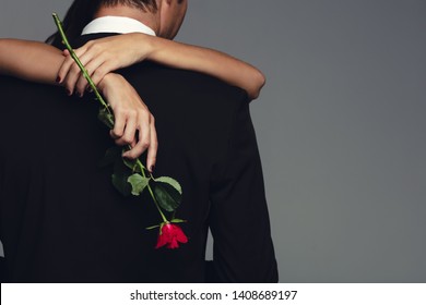 Rear View Of Woman Holding A Rose Embracing Man In Black Suit. Loving Couple Embracing On Grey Background.