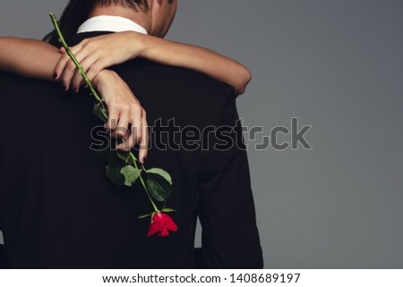 Rear view of woman holding a rose embracing man in black suit. Loving couple embracing on grey background.