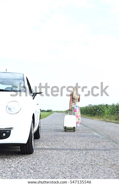 Rear view of woman with luggage leaving broken down\
car on country road
