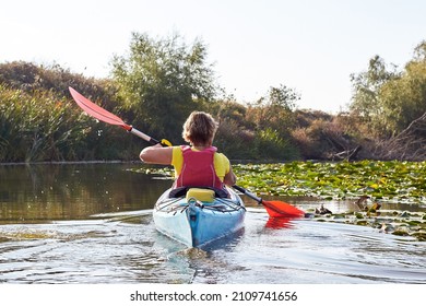 Rear view of woman paddle in blue kayak in the small river among the water lilies and driftwoods
