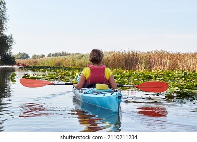 Rear view of woman paddle in blue kayak in river among the water lilies and driftwoods