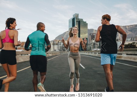 Rear view of woman running backwards with her friends on city street. Group of multi-ethnic friends training together outdoors in morning.