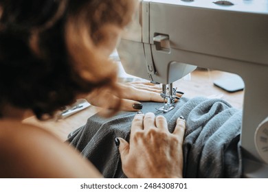 Rear view of a woman seamstress who sits at a sewing machine and sews summer pants from light gray fabric. The work of a professional tailor in an atelier. Tailoring of clothes to order. - Powered by Shutterstock