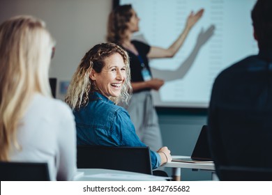 Rear View Of A Woman Sitting In Audience Looking Back At A Colleague And Smiling During A Conference. Businesswoman Smiling During A Presentation.