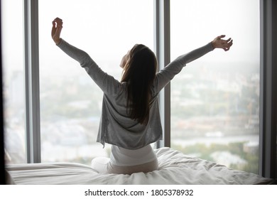 Rear view woman sitting on cozy bed in bedroom, stretching hands after awakening, dreamy young female enjoying morning, starting new day in modern apartment with large panoramic windows