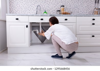 Rear View Of A Woman Trying To Stop Water Leakage From Sink Pipe In Kitchen