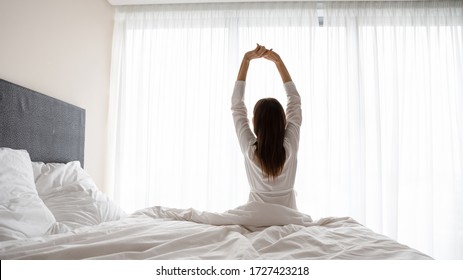 Rear view woman wake up sit in bed raised hands stretching body start day right with exercises, increases muscles tone encouraging joints flexibility, energy boost, enjoy new day, good morning concept