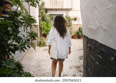 Rear view of a woman walking in a narrow alley with plants, wearing a white shirt and tote bag, enjoying a relaxed stroll. - Powered by Shutterstock