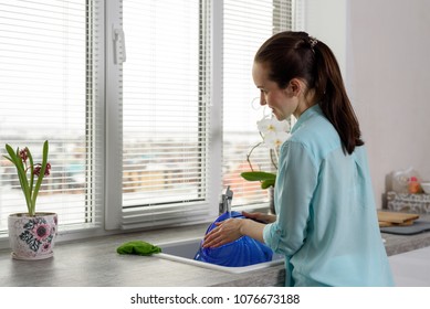 Rear View Of A Woman Washing Dishes In The Kitchen In Front Of The Window.