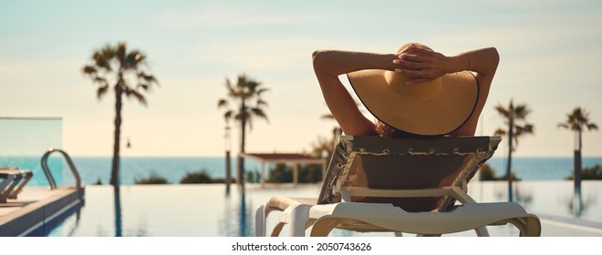 Rear view woman wear hat lying on deckchair near pool, put hands behind head relaxing, take sun bath, sea palm tree empty swimming pool scenery on background. Summer holidays, vacation, travel concept - Shutterstock ID 2050743626