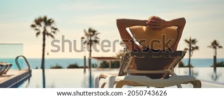 Rear view woman wear hat lying on deckchair near pool, put hands behind head relaxing, take sun bath, sea palm tree empty swimming pool scenery on background. Summer holidays, vacation, travel concept