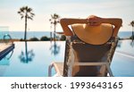 Rear view woman wear straw hat lying on deckchair on poolside put hands behind head relaxing take sun bathing, sea, palm tree, empty swimming pool scenery on background. Summer holidays travel concept