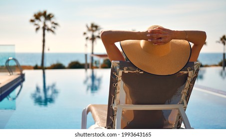 La mujer de la vista trasera lleva sombrero de paja en la tumbona junto a la piscina, pone manos detrás de la cabeza relajante tomar el sol, mar, palmera, paisaje vacío de la piscina en el fondo. Concepto de viajes de vacaciones de verano