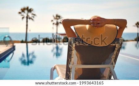 Rear view woman wear straw hat lying on deckchair on poolside put hands behind head relaxing take sun bathing, sea, palm tree, empty swimming pool scenery on background. Summer holidays travel concept