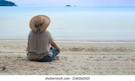 Rear view of woman wearing hat sitting on the sand looking at the calm ocean.  - Shutterstock ID 2160262425