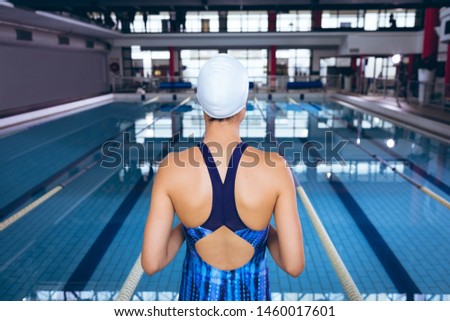 Rear view of a woman wearing a swimsuit and swimming cap standing by an olympic sized pool inside a stadium