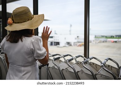 Rear View Of Women Wearing A Hat In The Airport Wave Goodbye Through The Window. On The Concept Of Plane Travel And Transportation