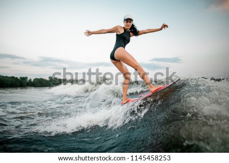 Rear view young active brunette girl riding on the wakesurf on high wave of motorboat on the background of trees