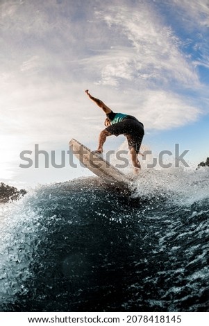 rear view of young active man balancing on wave on wakesurf board on splashing wave. Blue sky at the background.