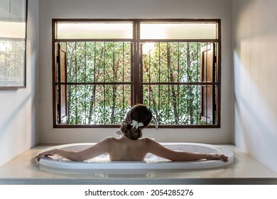 Rear view of a young adult woman with a flower on her hair she is taking a bath and lying in Bubbles foam water in the morning. She is Enjoying and relaxing in a spa hotel. Selective and soft focus.