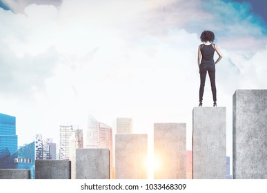 Rear view of a young African American businesswoman wearing a suit and high heels. She is standing on a giant bar chart and looking at a morning city. Mock up toned image - Shutterstock ID 1033468309