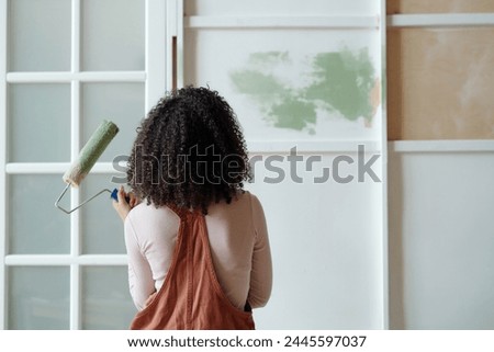 Rear view of young African American female owner of cafe with paintroller in hand standing in front of wall while painting it in green color