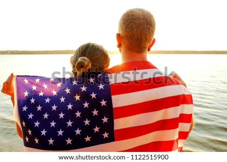 Rear view of young American patriotic couple hugging with USA flag on their backs enjoying beautiful summer sunset by the river. Independence day celebration concept. Background, close up, copy space.