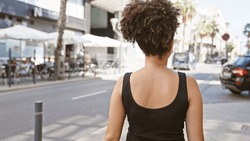 Rear View Of A Young Attractive Hispanic Woman With Curly Hair Walking On An Urban City Street During Daytime.