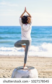 Rear View Of Young Black Woman Doing Yoga In The Beach. Female Wearing White Sport Clothes In Tree Asana With Defocused Sea Background. Afro Hairsytle.