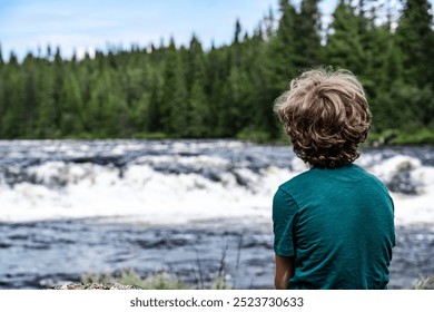 Rear view of a young blond boy sitting on rocky riverbank, observing the rushing rapids of Storån River, surrounded by a dense forest on a warm summer day in the Nordic Wilderness - Powered by Shutterstock